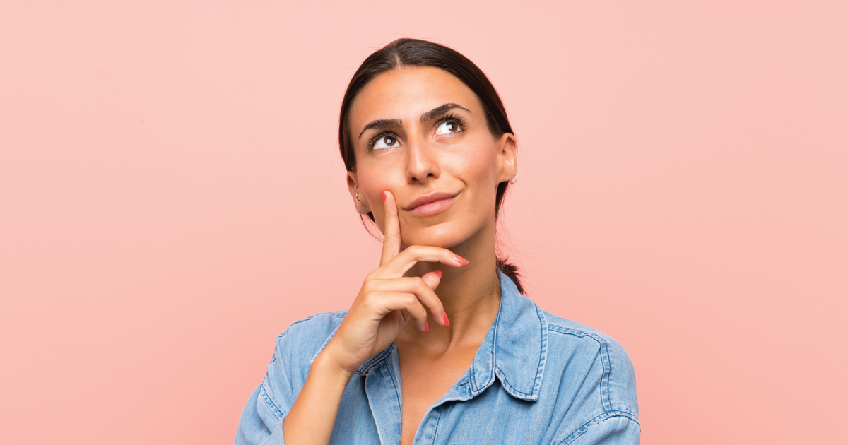 Donna pensierosa che guarda verso l’alto con il dito sul mento, indossa una camicia di jeans su sfondo rosa, simbolo di riflessione o curiosità.